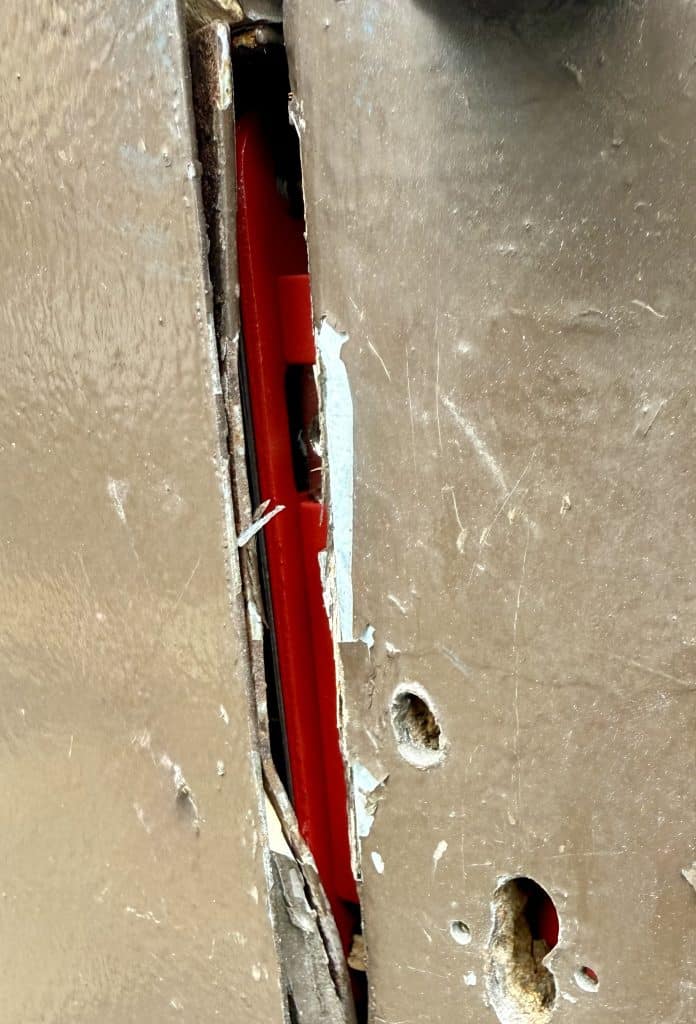 Close-up photo showing severe damage to the edge of a brown, painted wooden fire door (FD60). A large, ragged keyhole has been cut out of the door where a lock was incorrectly installed. The wood is splintered and split, and a red sash lock in place of a deadlock no intumescent fire safety strip is visible in the gap between the door and the frame, which has been compromised by the poor installation this is a consequence of calling poorly trained locksmiths.